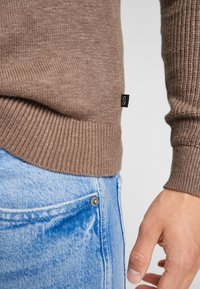 Brauner Strickpullover mit geripptem Saum, kombiniert mit hellblauen Jeans. Ein kleines schwarzes Etikett ist an der Seite des Pullovers sichtbar.