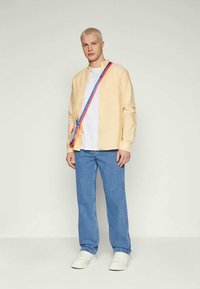 Lichtgele button-up shirt, wit T-shirt eronder, blauwe jeans, witte sneakers en een gekleurde schoudertas. Eenvoudig, casual ontwerp.
