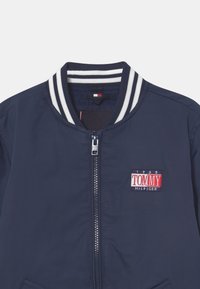 Marine bomberjack met wit gestreepte geribde kraag, ritssluiting en geborduurd "TOMMY HILFIGER" logo in rood en blauw. Glad nylon materiaal.