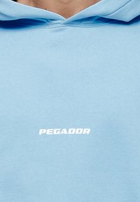 Sudadera con capucha azul claro hecha de material suave con un bolsillo tipo canguro en la parte frontal. Presenta un logo blanco "PEGADOR" en el centro.