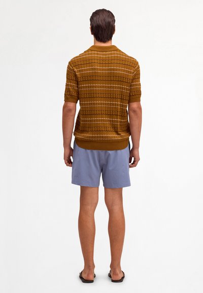 Man wearing brown striped short-sleeve knit shirt, light blue shorts, and black sandals standing facing away against white background.