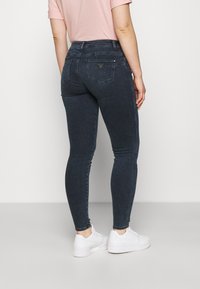 Persona vestida con jeans ajustados de color azul oscuro, una camiseta de manga corta rosa clara y zapatillas blancas de pie sobre un fondo liso claro.