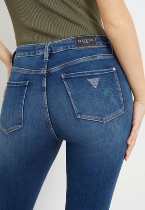Primo piano di una persona che indossa jeans Guess blu con il logo triangolare di strass sulla tasca posteriore e un top a coste verde oliva.