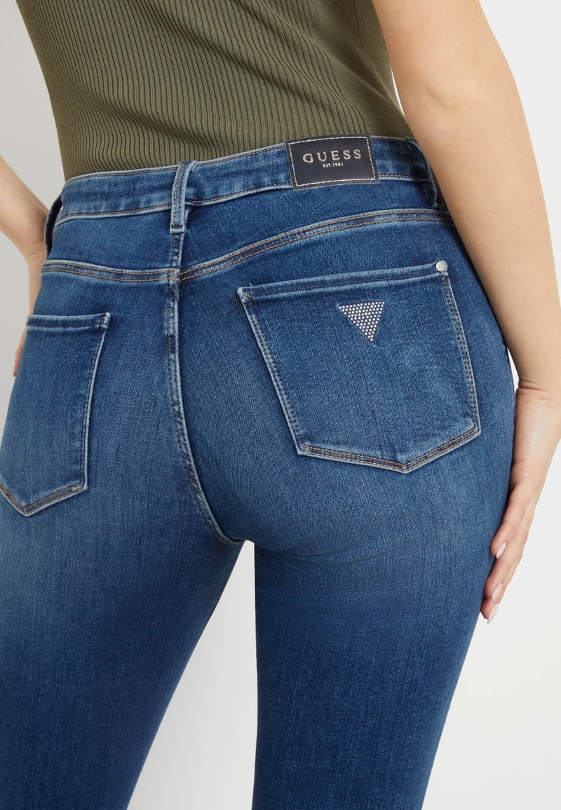 Gros plan d'une personne portant un jean Guess bleu avec le logo triangulaire en strass sur la poche arrière et un haut côtelé vert olive.