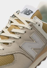 Μπεζ και μουσταρδί αθλητικό παπούτσι με λευκά κορδόνια και μεγάλο λευκό λογότυπο "N" στο πλάι, με λεπτομερή ραφές και ορατό λογότυπο της μάρκας στη γλώσσα.