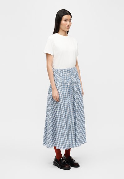 Γυναίκα που στέκεται, φοράει λευκό τοπ με κοντά μανίκια, μπλε και λευκή καρό μίντι φούστα, σοκολατί κάλτσες και μαύρα μοκασίνια.