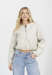 Veste courte gris clair avec un col rond, des manches longues et une fermeture éclair à l'avant. Associée à un jean bleu taille haute. Texture lisse.