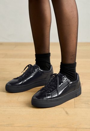 Beneden in zwarte doorzichtige panty's en sokken, met glanzende zwarte platformsneakers met zijritsen, op een houten vloer.