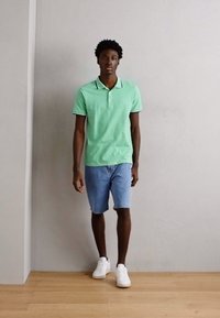 Camisa de polo de algodón verde de mangas cortas, combinada con pantalones cortos de mezclilla azul claro. Terminada con zapatillas blancas sobre un suelo de madera.