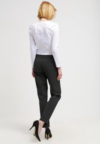 Chemise blanche à manches longues, cintrée à la taille, associée à un pantalon noir ajusté et des escarpins noirs. Fond uni.