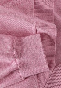 Ružičasta pletena vestica, s rebrastim rukavima i mekanom, teksturiranom površinom. Detalji uključuju suptilni heather uzorak.