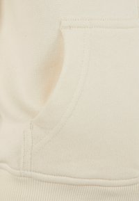 Tissu de sweatshirt beige clair avec une poche kangourou, un ourlet côtelé et des détails cousus. Texture lisse avec une coupe ajustée.