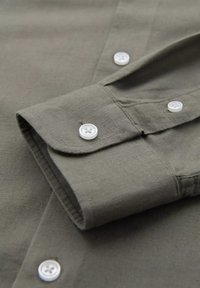 Una camicia di lino verde oliva con maniche e polsini abbottonati, caratterizzata da bottoni bianchi e una sottile texture che mette in risalto la sua qualità e maestria artigianale.