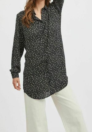 Kobieta ubrana w długą czarną bluzkę na guziki z białym kwiatowym wzorem i luźne kremowe spodnie, podnosząca jedną rękę na jednolitym tle.