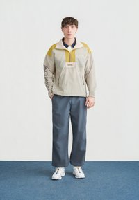 Άνδρας που στέκεται σε μπλε χαλί, φορόντας μπεζ και κίτρινο μπουφάν Columbia, μπλε παντελόνι με φαρδιά μπατζάκια και λευκά αθλητικά παπούτσια με χρωματικές λεπτομέρειες.