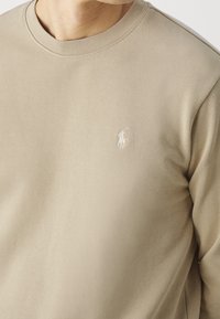 Beige bomullströja med rund hals. Har en liten vit broderad logotyp p å vänster bröst. Mjuk textur, avslappnad passform.