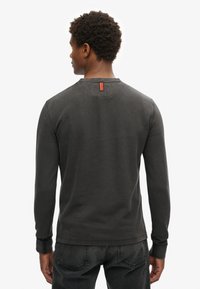 Mężczyzna z krótkimi kręconymi włosami, ubrany w ciemnoszarą koszulę z długim rękawem i czarne dżinsy, stoi tyłem na białym tle.