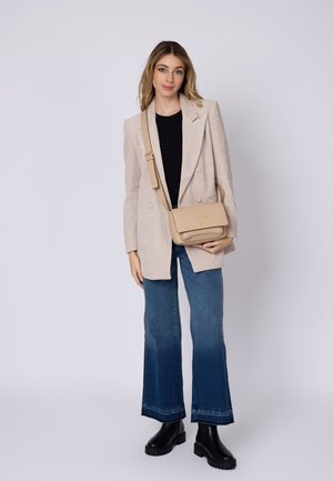 Beige läder crossbody-väska med en lock, prydd med en guld-logotyp, bärs över en ljusbeige kavaj, ihop med utsvängda blå jeans och svarta stövlar.