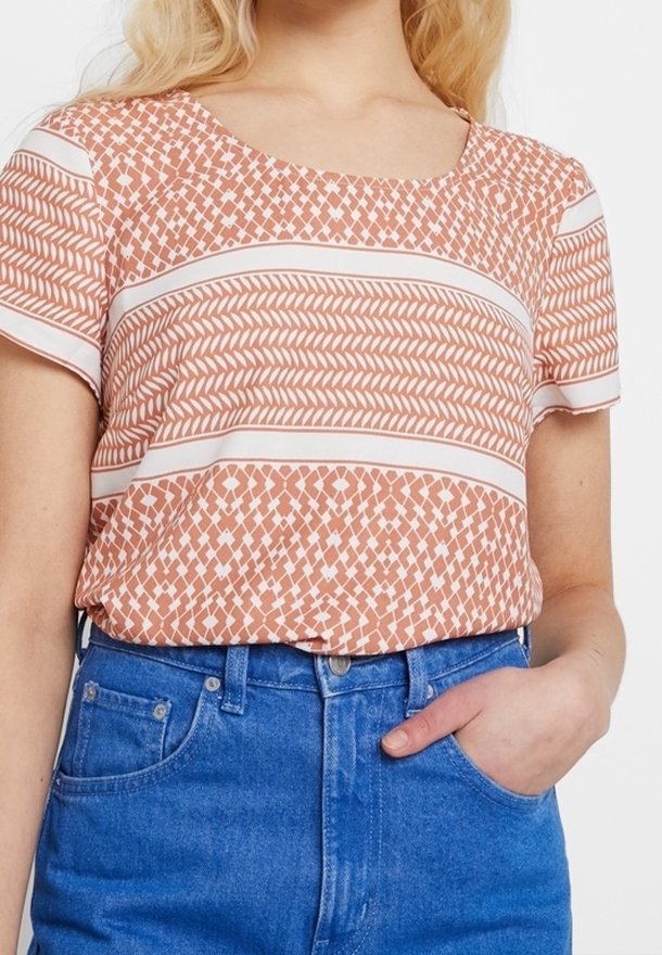 Femme portant un haut à manches courtes à motif pêche et blanc, rentré dans un jean bleu taille haute, avec une main dans une poche.