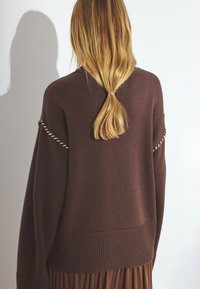 Brun stickad tröja med ribbad nederkant, med kontrasterande sömmar på axlarna. Texturerad tyg, avslappnad passform och mittlång design.