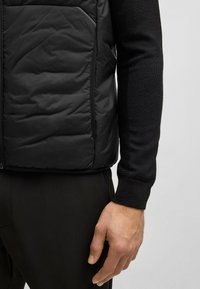 Schwarze, isolierte Weste mit gestepptem Muster und Seitentaschen, getragen zu einem taillierten schwarzen Langarmshirt und dunklen Hosen. Wollbündchen.