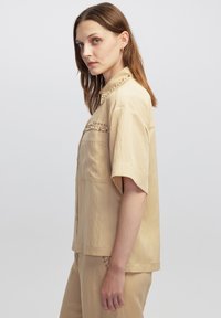 Camisa beige de manga corta con cierre de botón, que presenta tachuelas decorativas a lo largo del cuello y el bolsillo del pecho, fabricada con una tela ligera.