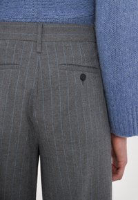 Pantaloni grigio a righe sottili in lana, con una tasca posteriore dotata di un bottone nero e una texture morbida, abbinati a un maglione di maglia azzurro chiaro.