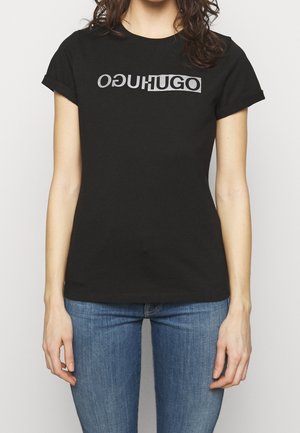 Musta puuvillat-paita, jossa on rohkea, heijastava logo "OḠUHUGO" rinnassa; lyhyet hihat ja istuva muotoilu.