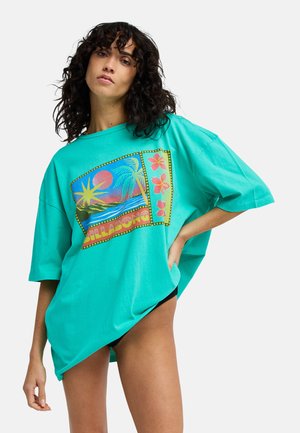 Kobieta z kręconymi włosami ubrana w oversize turkusowy t-shirt Billabong z tropikalnym zachodem słońca i kwiatowym motywem, stojąca z ręką na biodrze.