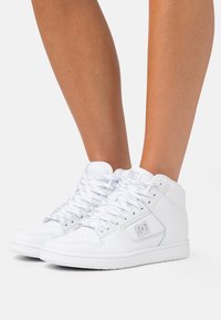 Hvite høye joggesko laget av syntetisk materiale med flate lisser, perforert tå og en teksturert gummisåle; logo på siden.