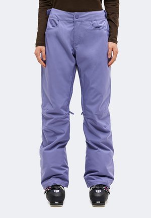 Une personne portant un pantalon de ski ample couleur lavande avec des poches et des chaussures de ski noires, debout devant un fond uni.