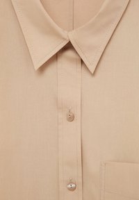 Chemise beige à col classique avec des boutons, dotée de coutures verticales et d'une poche sur le côté gauche. Tissu lisse avec un léger éclat.