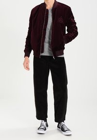 Bomber jacket in velluto bordeaux con polsini a costina e logo, indossato sopra una maglietta a girocollo grigia chiara, abbinato a pantaloni in corduroy neri e sneakers.