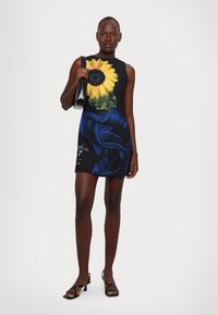 Black sleeveless mini dress featuring a large sunflower graphic in vibrant yellow, with blue floral accents and smooth texture. Holding a black handbag.