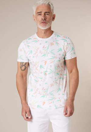 Homme d'âge moyen aux cheveux blancs et à la barbe portant un t-shirt blanc à motif floral et un pantalon blanc, debout devant un fond neutre.