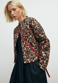Jakke med leopardmønster i nuancer af orange og sort, med et slankt design og en rød skulderrem. Bæres over en sort kjole.