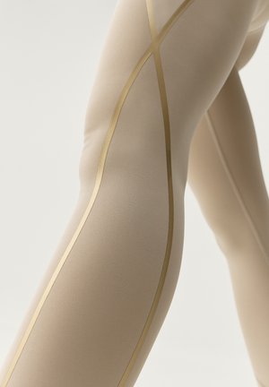 Leggings beige con una textura suave y líneas de acento doradas que crean un patrón en X a lo largo del costado. Vista en primer plano de la tela.
