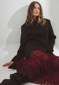 Frau mit welligem Haar, die an einer weißen Wand lehnt, in einem schwarzen Blazer, einem burgunderfarbenen Spitzenrock und schwarzen High-Heel-Stiefeln, sitzt auf dem Boden.