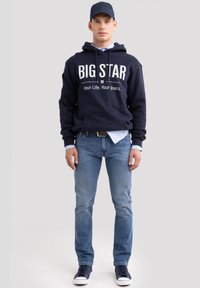 Jaunas vyras dėvi tamsiai mėlynę kepurę, tamsiai mėlyną „BIG STAR“ džemperį su gobtuvu, šviesiai mėlyną marškinius, mėlynas džinsus ir tamsiai mėlynas sportbačius, stovintis priešais paprastą foną.