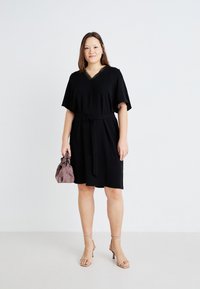 Black dress with a V-neck, short sleeves, and a fabric belt, featuring a textured neckline. Worn with beige heeled sandals and a lavender bag.