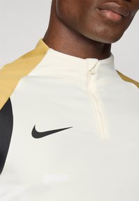 Nike zip-up majica s kremastom tkaninom i žutim i crnim detaljima. Dizajn uključuje ovratnik s polu-razipom i istaknuti logotip na prsima.