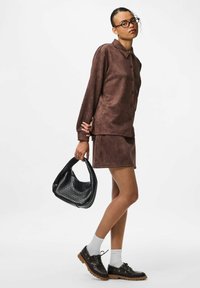 Brun ruskinds skjorte med knapper og matchende nederdel, parret med en sort vævet håndtaske, hvide sokker og mørke sko med en kraftig sål.