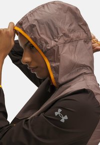 Lightweight hooded jacket in brown with orange accents; features a textured finish and a logo on the sleeve. Adjustable hood for fit.