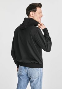 Czarna kurtka z kapturem o gładkiej fakturze, luźnym kroju i prążkowanych mankietach, zestawiona z jasnoniebieskimi, przecieranymi dżinsami.