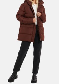 Femme portant une doudoune matelassée marron à capuche, un haut blanc, un jean noir et des bottines noires, debout avec une jambe légèrement pliée.