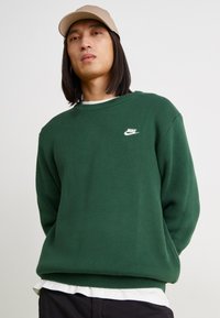 Πράσινη πλεκτή μπλούζα με στρογγυλή λαιμόκοψη και ραμμένα μανίκια, με μικρό λευκό λογότυπο Nike στο στήθος. Υφασμάτινη υφή, χαλαρή εφαρμογή.