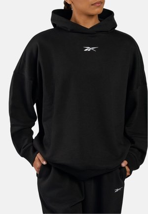 Personne portant un sweat à capuche noir Reebok et un pantalon de survêtement noir assorti avec des logos blancs Reebok, mains dans les poches, légèrement tournée vers la droite.
