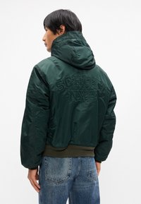 Zelena bomber jakna s kapuljačom, s podstavljenom teksturom, s vezom "SCHOTT NYC" na leđima i rebrastim detaljima na donjem rubu.