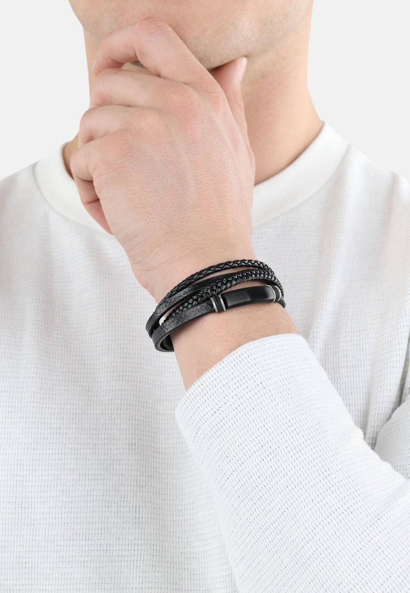 Braccialetto in pelle nera con tre fili intrecciati; superfici texturizzate e lisce. Indossato su un polso con una camicia bianca a maniche lunghe.