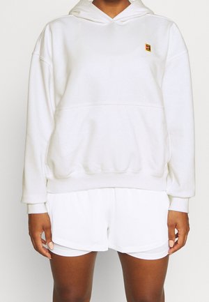 Personne portant un sweat à capuche blanc ample avec un petit logo jaune et rouge sur la poitrine et un short blanc superposé, debout devant un fond uni.